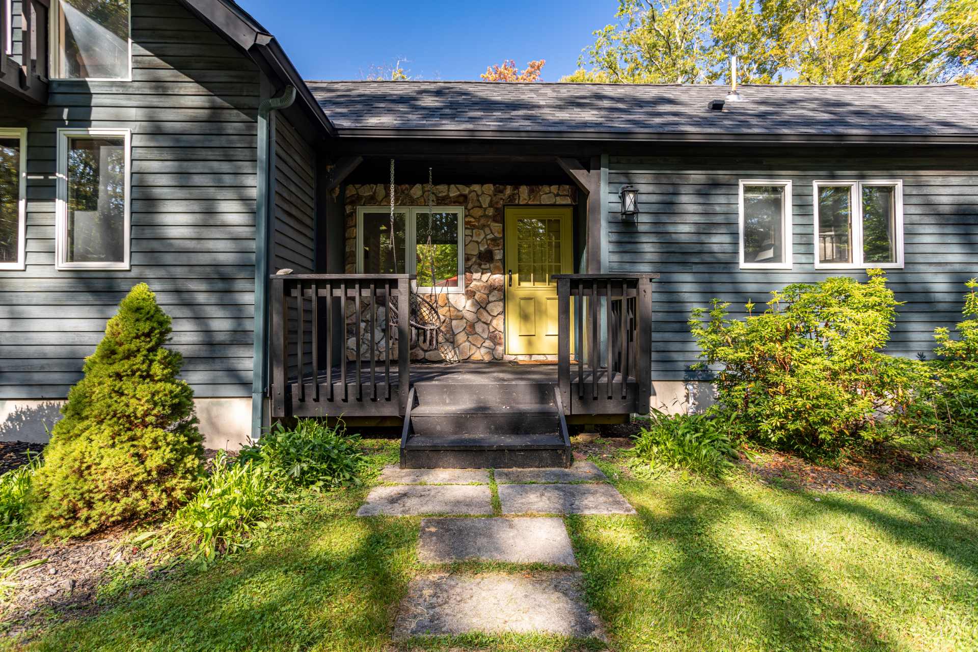 The front entrance of the dark-sided house features a small covered porch with a yellow door and stone accents. A wooden railing and steps lead up to the entrance, surrounded by greenery.