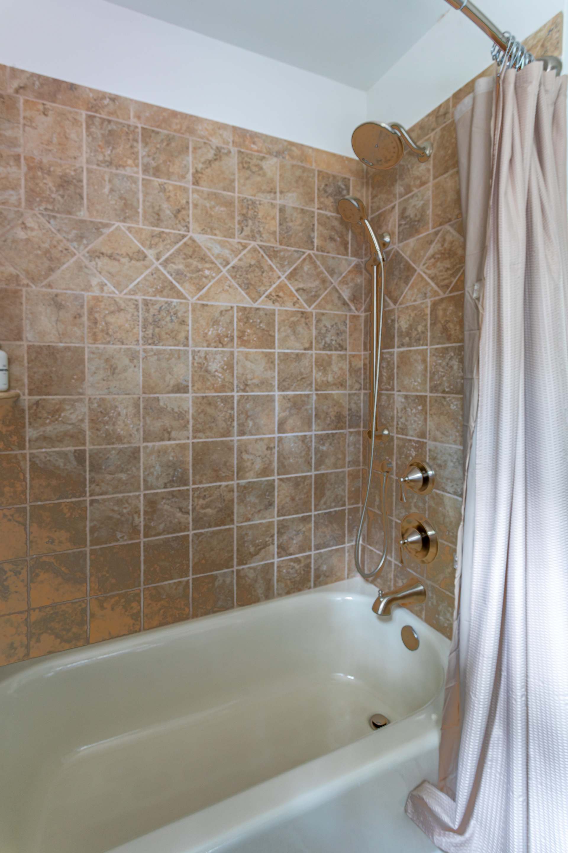 Close-up of a bathtub with light brown square tiles on the walls, featuring a shower head and a handheld sprayer. A light-colored shower curtain is pulled to the side.