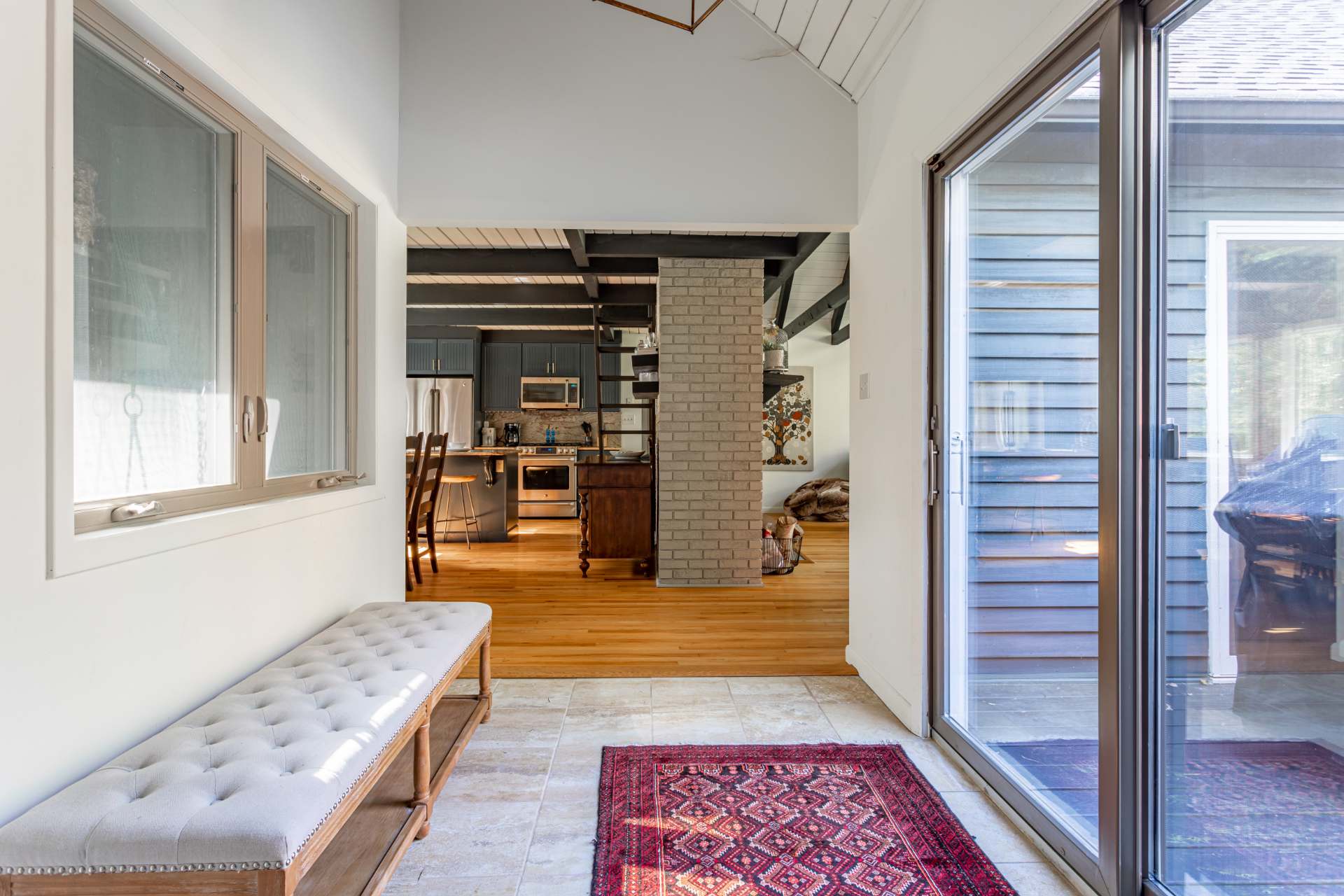 Entryway or foyer with tiled flooring, a long upholstered bench, and a red patterned rug. A sliding glass door leads to the exterior, and a view into the kitchen and living area is visible.