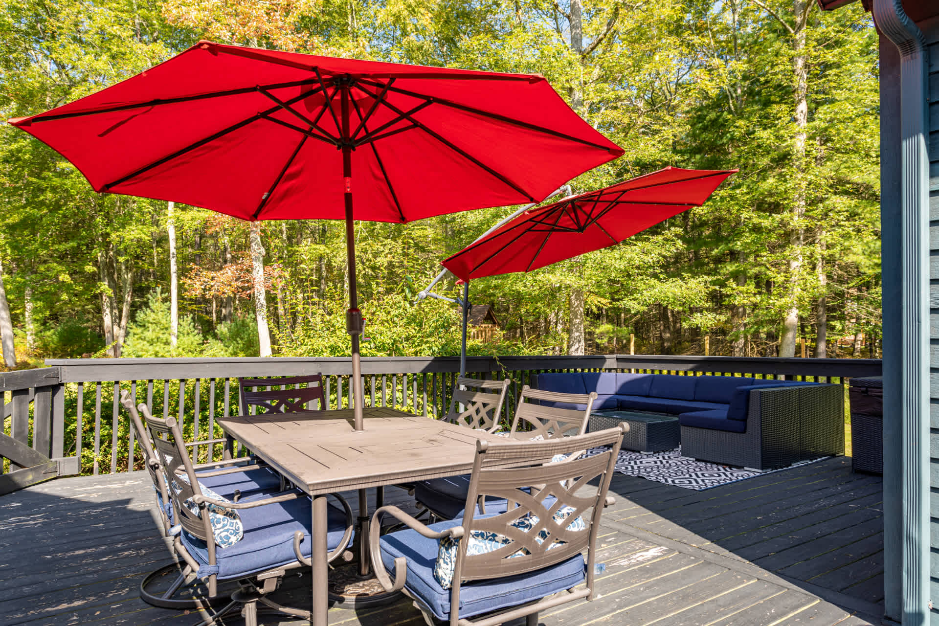 Expansive outdoor deck with dark wood planks, featuring a dining table with chairs under two large red umbrellas and a separate outdoor sofa seating area. Lush green trees provide a private, natural backdrop.