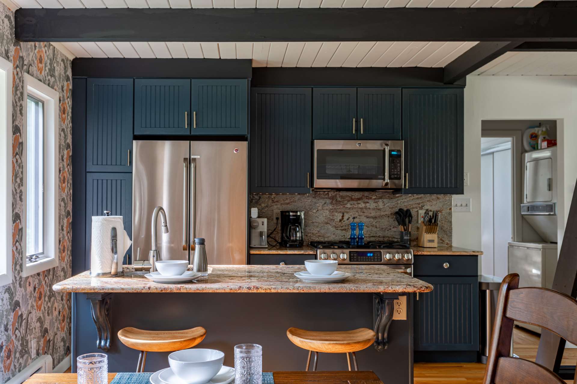 Open-concept kitchen with dark blue shaker-style cabinets, stainless steel appliances, granite countertops, and a breakfast bar with two wooden stools, adjacent to the dining area.