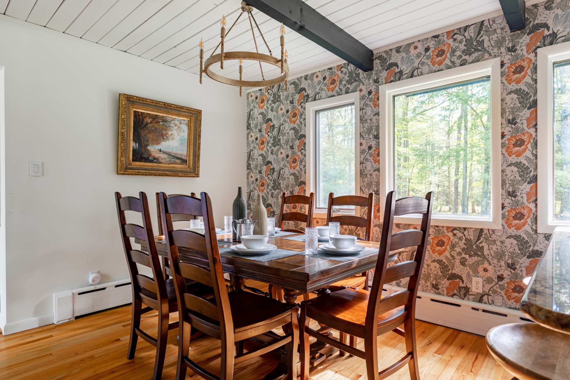 Dining room with a dark wooden rectangular table set for six, surrounded by matching chairs, featuring a striking floral wallpaper accent wall and a rustic circular chandelier.