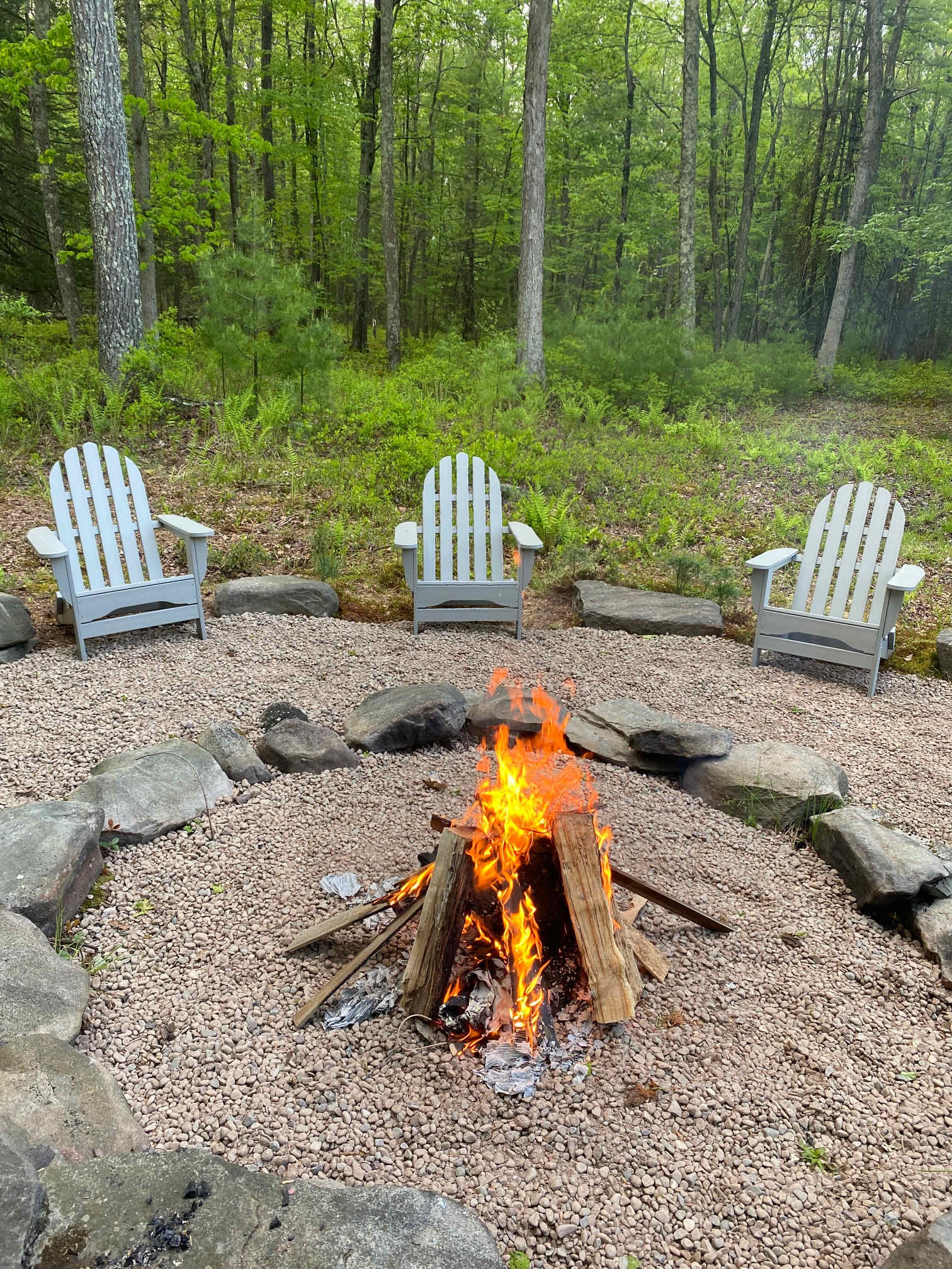 Outdoor fire pit area with a gravel base and large stone border, surrounded by three light gray Adirondack chairs. A crackling fire is lit, set against a backdrop of dense green forest.