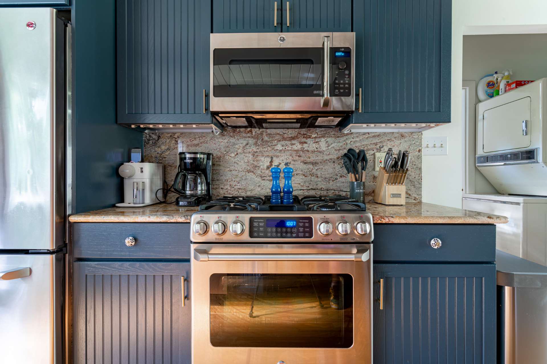 Close-up of the kitchen showing a stainless steel range with a microwave above, dark blue shaker-style cabinets, granite backsplash, and a coffee maker on the counter.