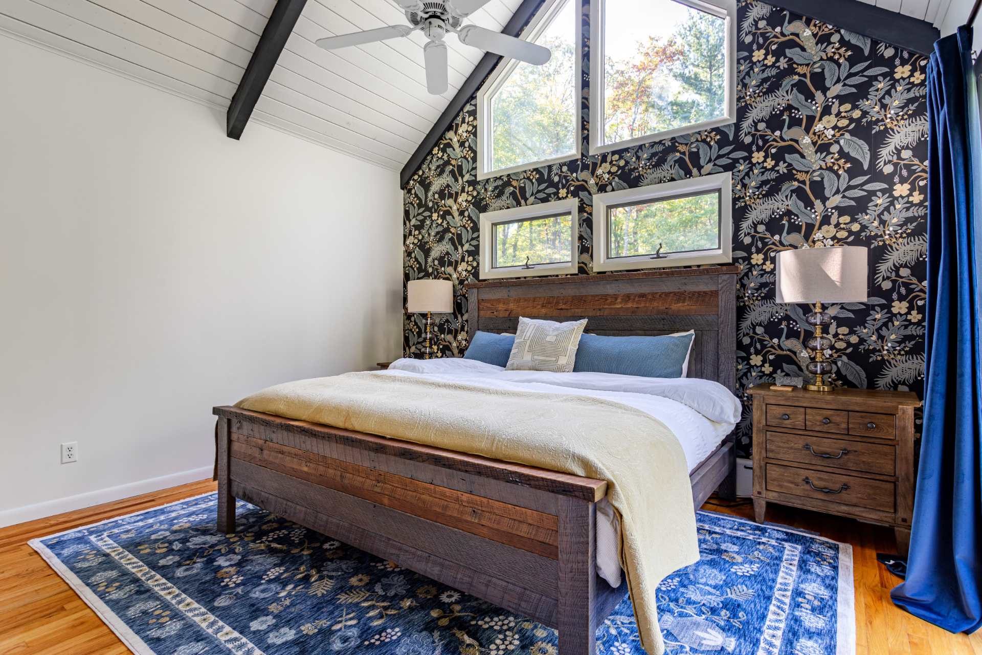 Master bedroom featuring a rustic wooden king-sized bed, a dark floral accent wall, vaulted ceiling with a fan, and high windows offering views of the surrounding trees.
