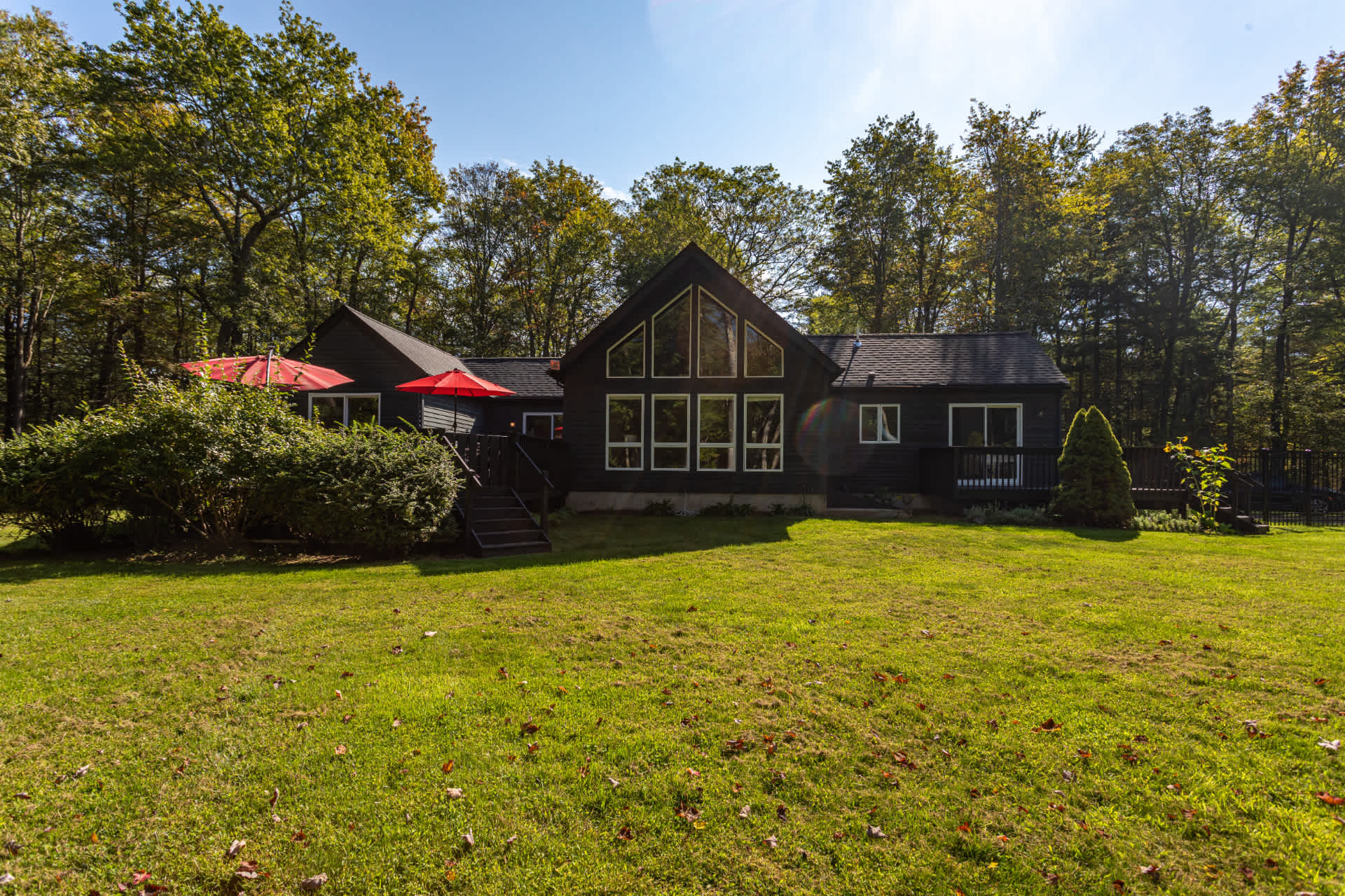 The back exterior of a dark-sided house with large windows and a deck is visible from a spacious green lawn. Red umbrellas and lush trees surround the property under a bright sky.