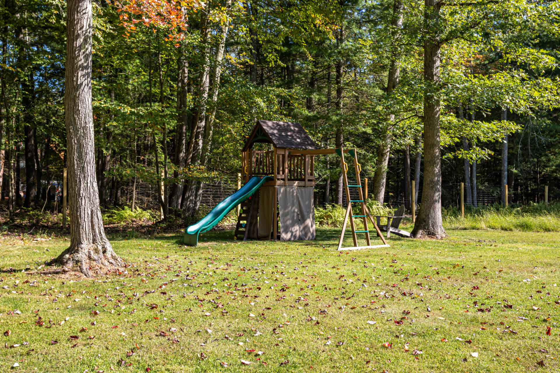 A wooden playset with a green slide and climbing frame is situated on a vibrant green lawn, framed by tall trees. Scattered leaves are visible on the grass, indicating an autumn setting.