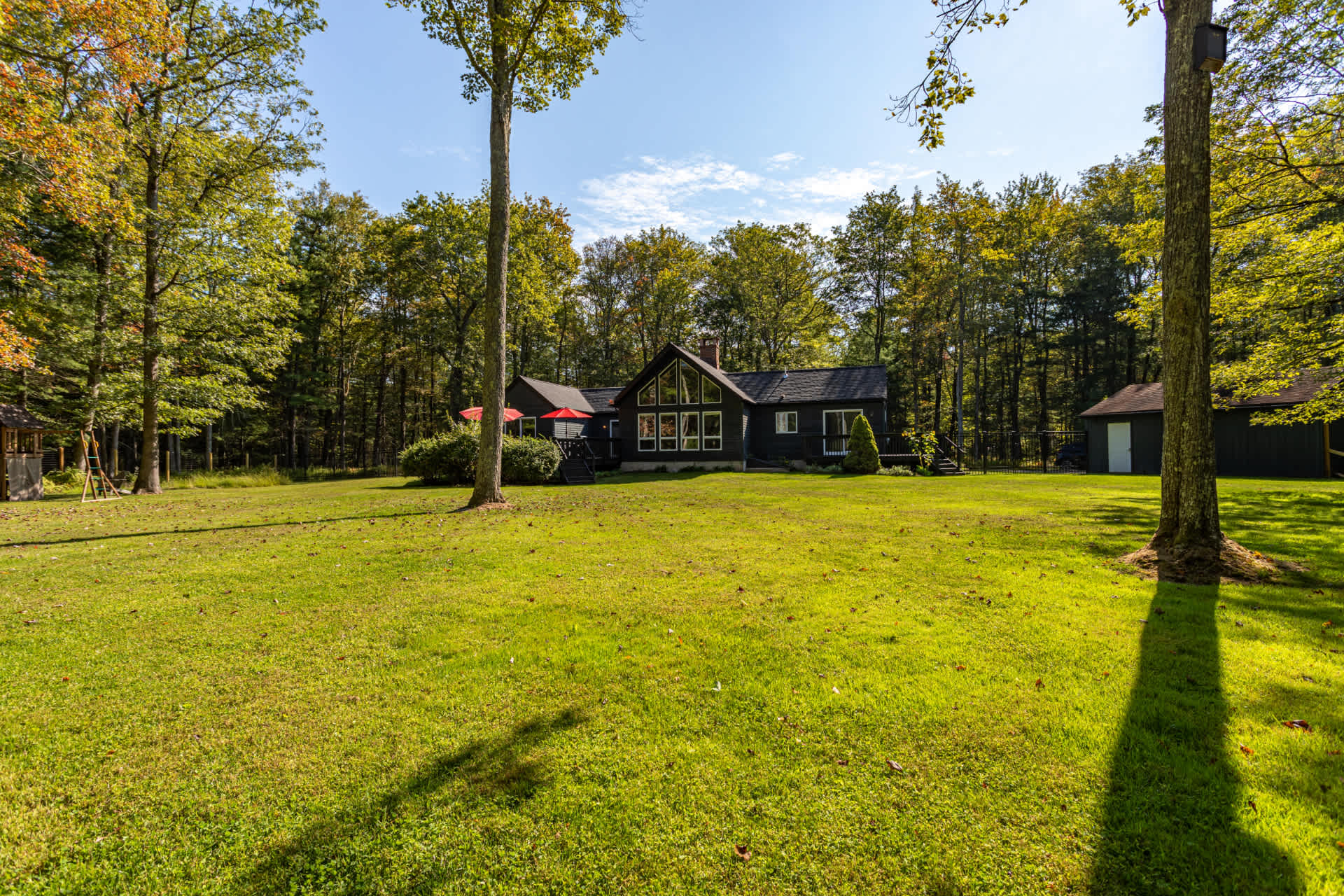 A wide shot of the dark-sided house from the expansive green backyard, surrounded by a dense forest. The house features large windows and a deck, with a small shed visible on the right.