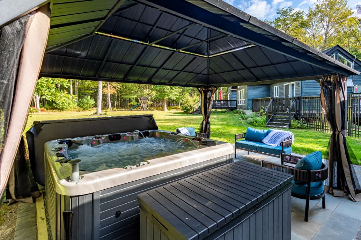 A bubbling hot tub is positioned under a dark metal gazebo, with blue outdoor cushions on nearby seating. The hot tub cover is partially open, revealing the water, and a dark storage box is next to it.