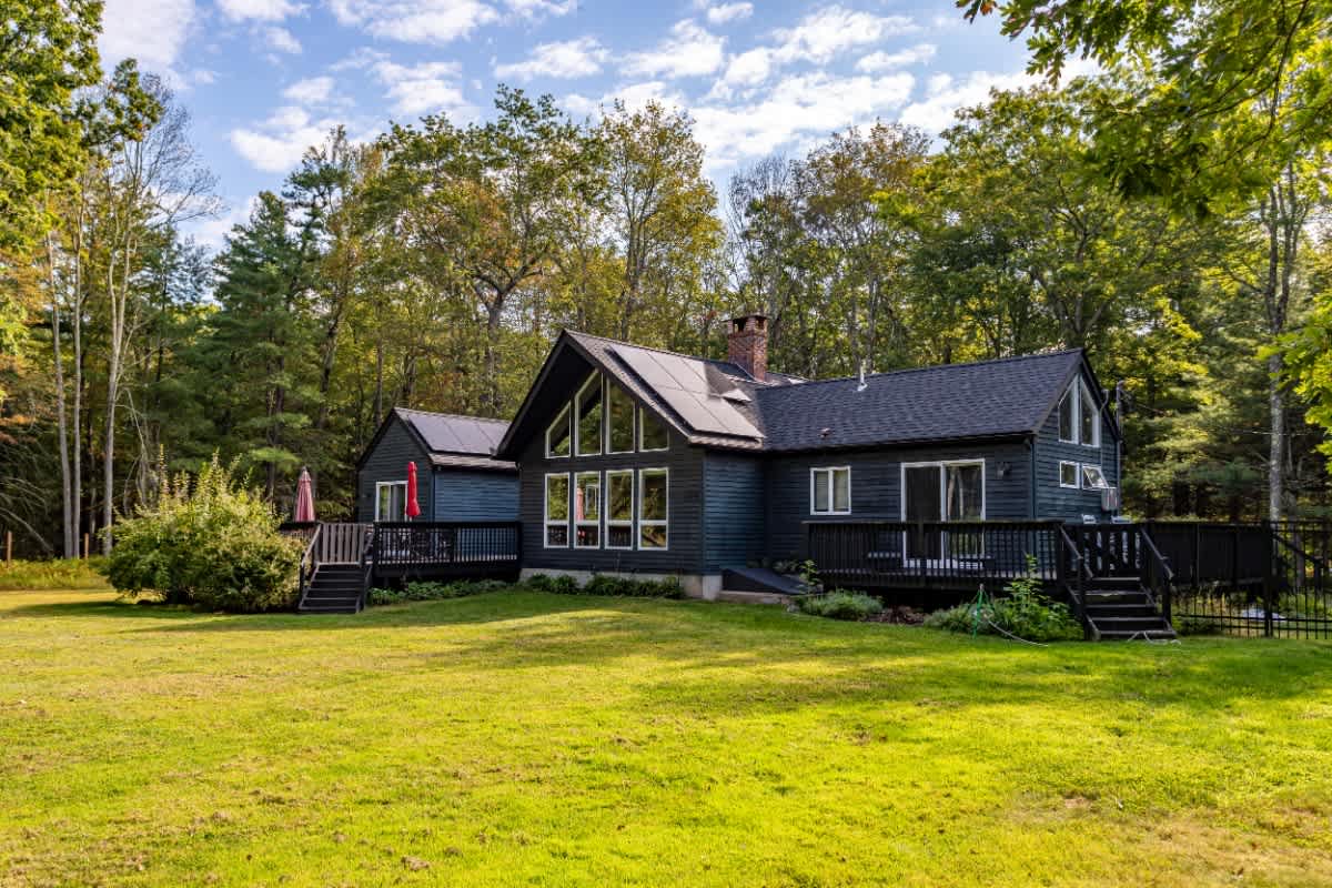 The modern dark-sided house features large windows, a prominent A-frame section, and solar panels on the roof, with a spacious wooden deck and stairs leading to a sprawling green lawn surrounded by mature trees.