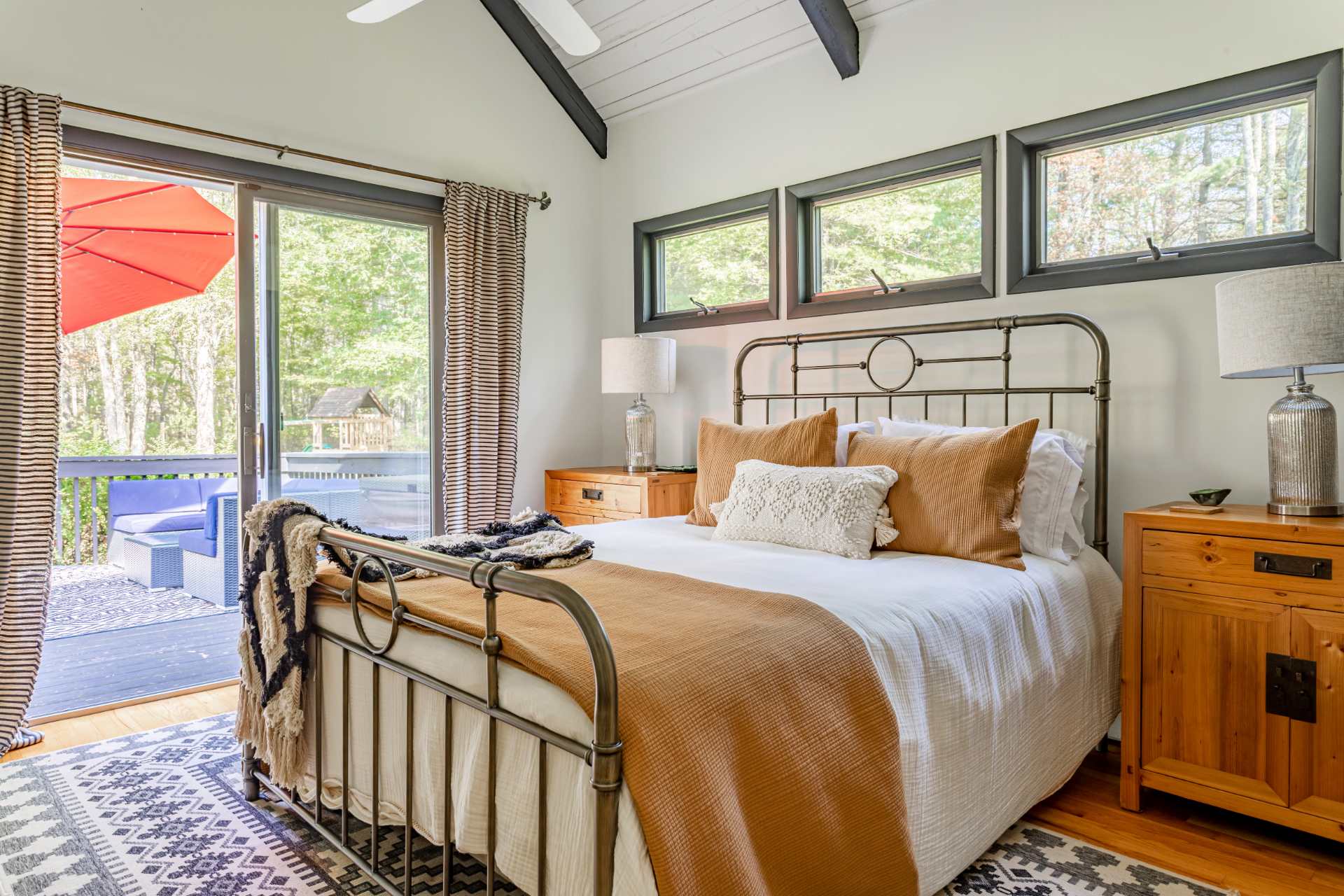 Master bedroom with a metal frame bed, white and rust-colored bedding, wooden nightstands, and a sliding glass door leading to a deck. Three small windows are above the bed, and the ceiling has exposed dark beams.