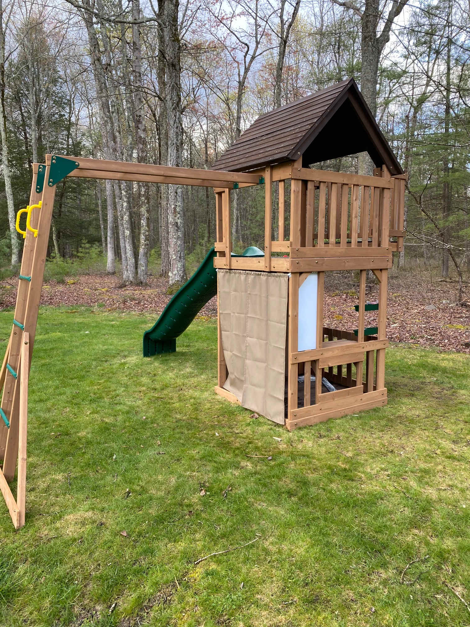 A wooden playset with a slide, climbing ladder, and small house structure is set on a green lawn, surrounded by a dense forest. The playset features a brown roof and a tan fabric enclosure.