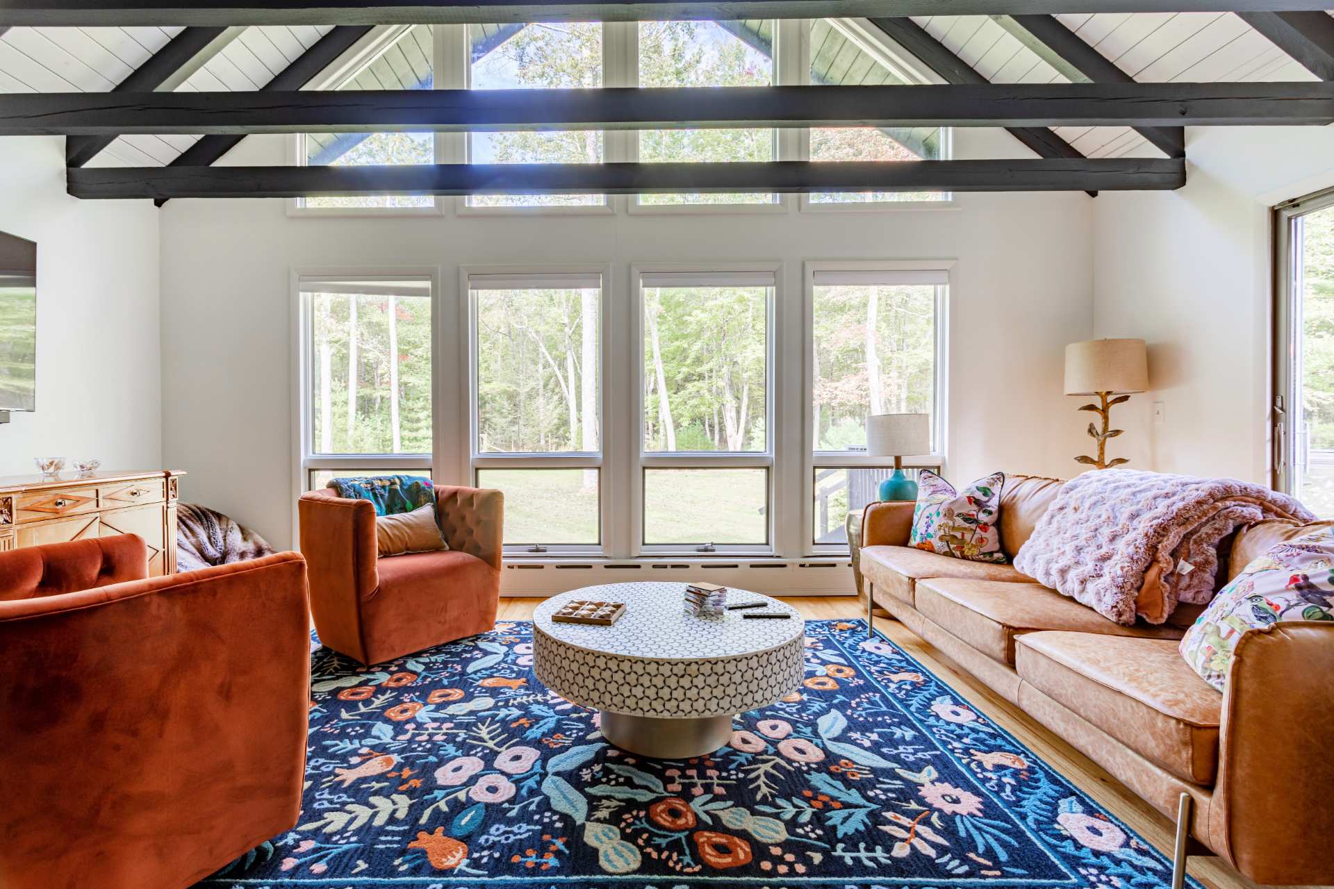 Bright living room featuring a high vaulted ceiling with exposed dark beams, large windows overlooking a wooded area, a brown leather sofa, two orange armchairs, and a vibrant blue floral rug.
