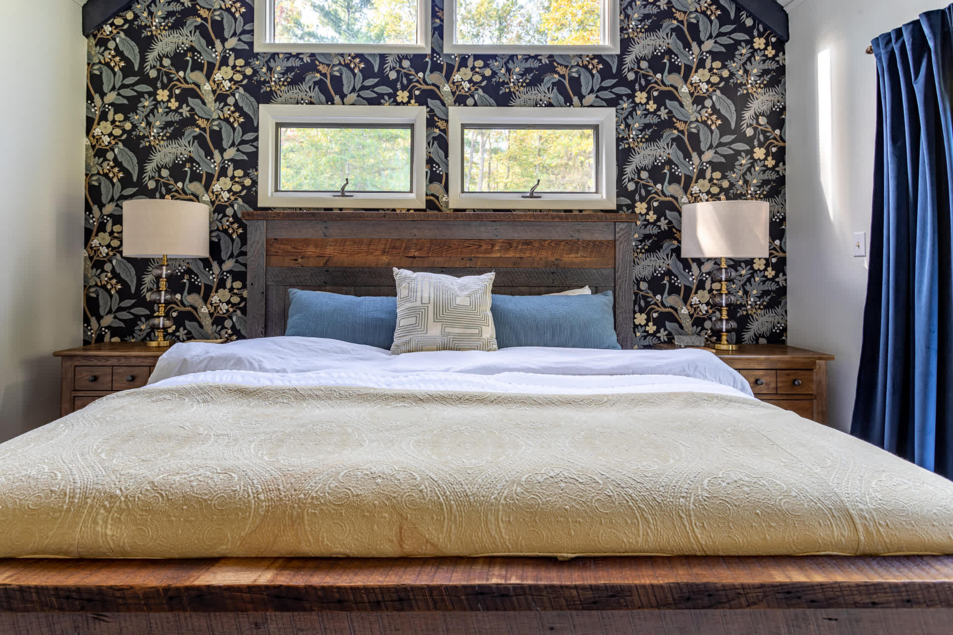 Close-up of the rustic wooden king bed in the master bedroom, highlighting the dark floral wallpaper, two nightstands with lamps, and small windows above the headboard.