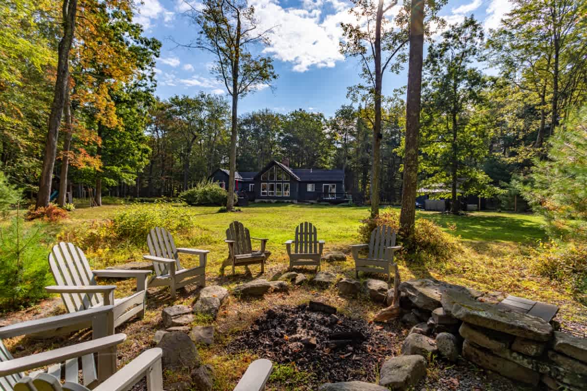 A cozy outdoor fire pit area featuring a stone-lined fire pit surrounded by several light-colored Adirondack chairs, set on a grassy lawn with tall trees and a dark modern house in the background.