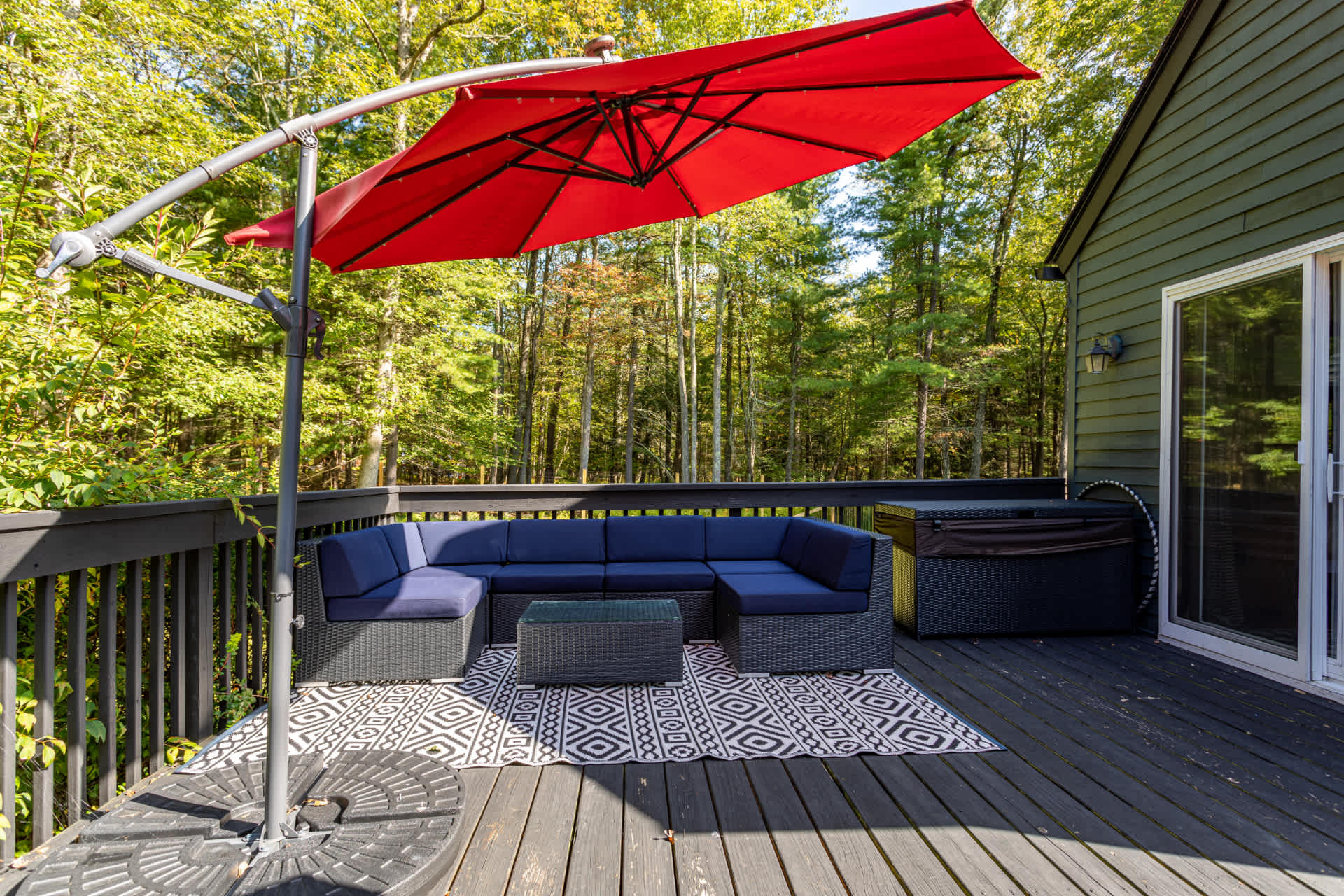 A dark wooden deck features a large blue sectional sofa with a glass-top coffee table, placed on a black and white geometric rug, shaded by a cantilevered red umbrella. A dark house exterior and forest are visible.
