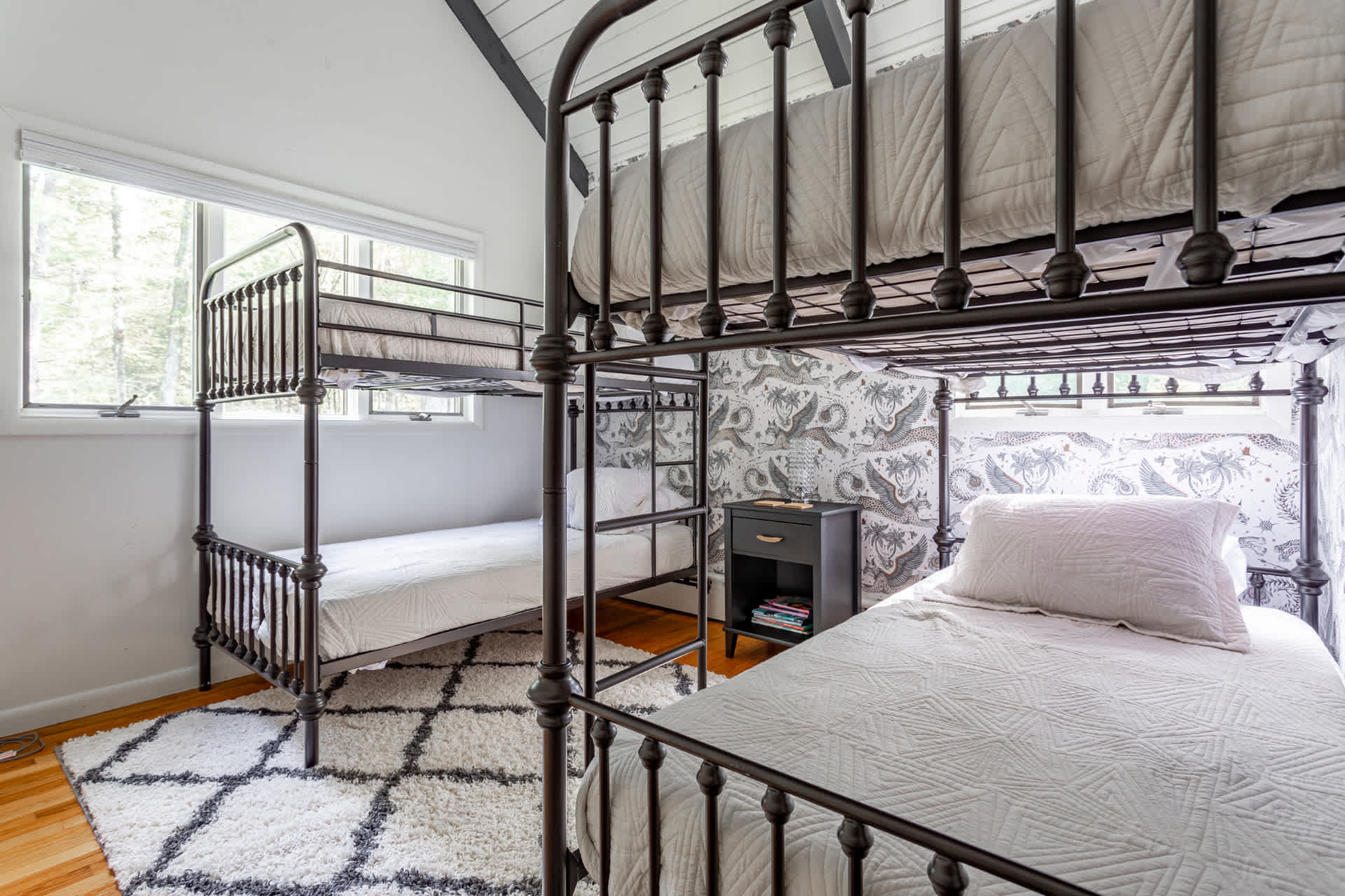 Kids' bedroom showing two black metal bunk beds with white bedding, a dark nightstand, and patterned wallpaper. A large window provides natural light, and a shaggy area rug is on the hardwood floor.