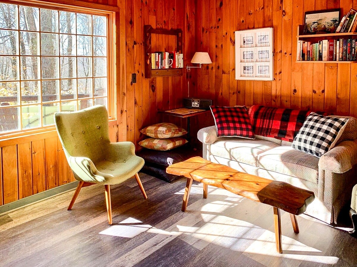 Bright living room with extensive wood paneling, a large window offering natural light and outdoor views, furnished with a light sofa, a green armchair, and a rustic wooden bench serving as a coffee table.
