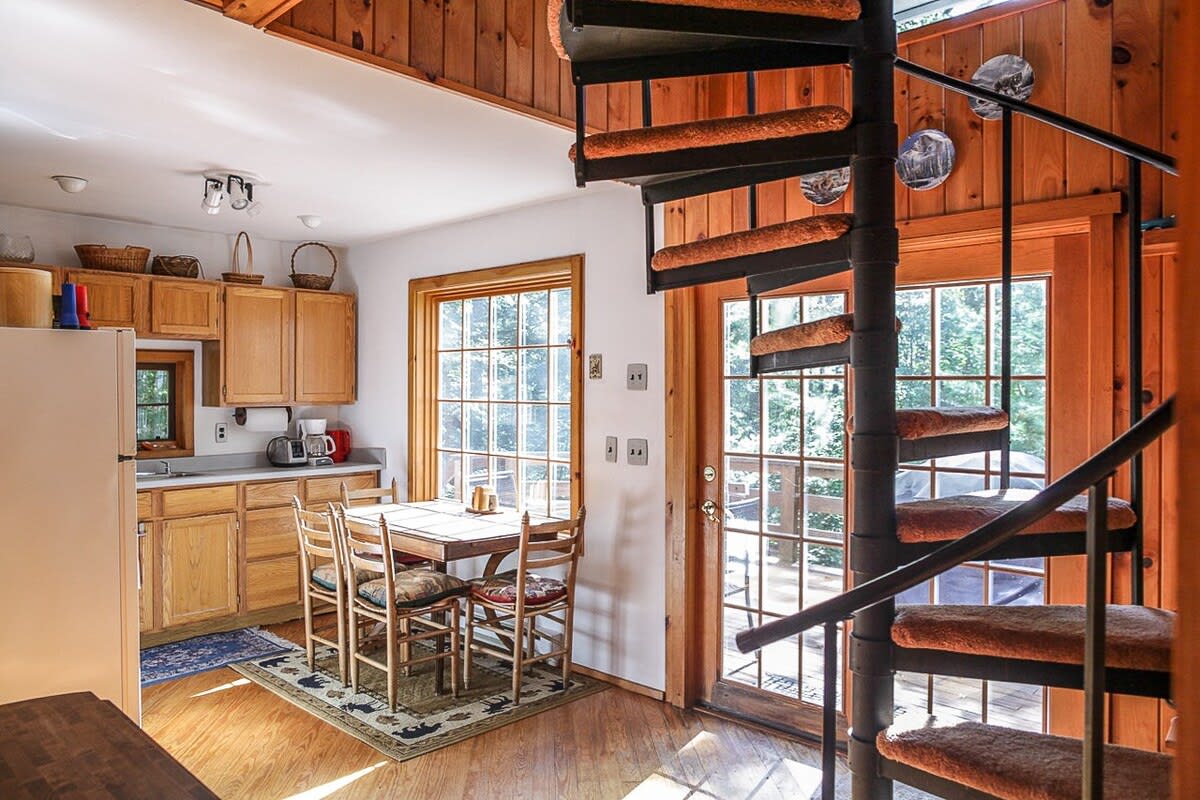 Open-plan kitchen and dining area with light wood cabinetry and a dining table, adjacent to a black spiral staircase with carpeted treads, leading to an upper level, with a glass door to the deck.