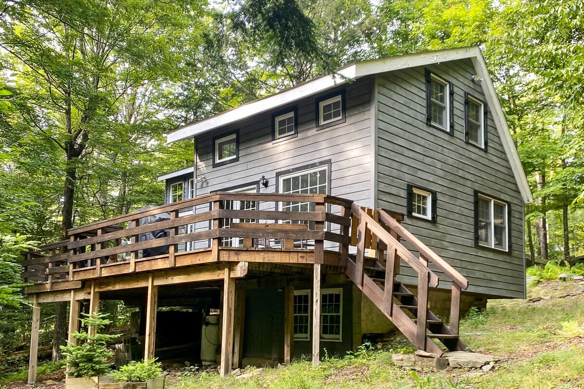 The back view of a gray-sided cabin with white trim and multiple windows, featuring a large wooden deck with stairs leading down to a grassy, wooded area. The cabin is surrounded by dense green trees.