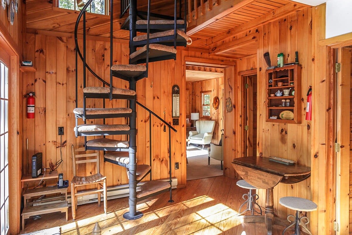 The entryway features warm wood-paneled walls and a prominent black spiral staircase with carpeted steps, leading to the upper floor, with a small dining table and a view into the living area.