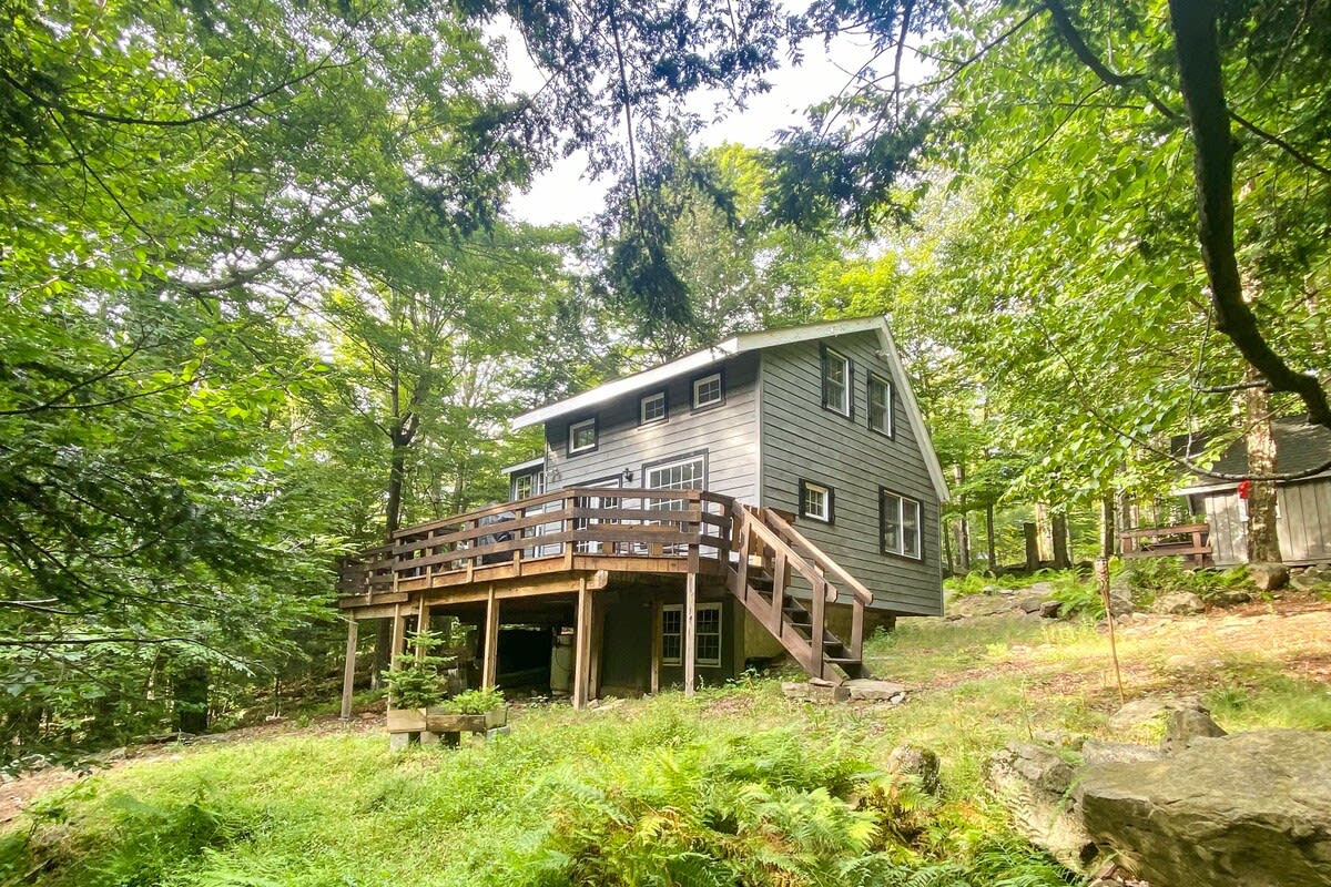A wider shot of the gray-sided cabin nestled in a lush green forest, showcasing its elevated wooden deck with stairs and the surrounding natural landscape. The property sits on a gentle slope amidst tall trees.
