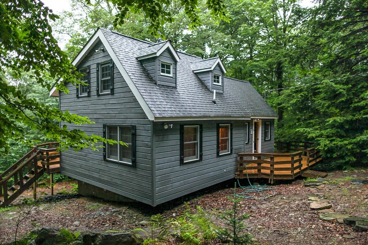 The gray-sided cabin stands on a sloped, wooded lot, featuring multiple windows with black shutters, a gabled roof with dormers, and a small wooden deck with stairs on the side.
