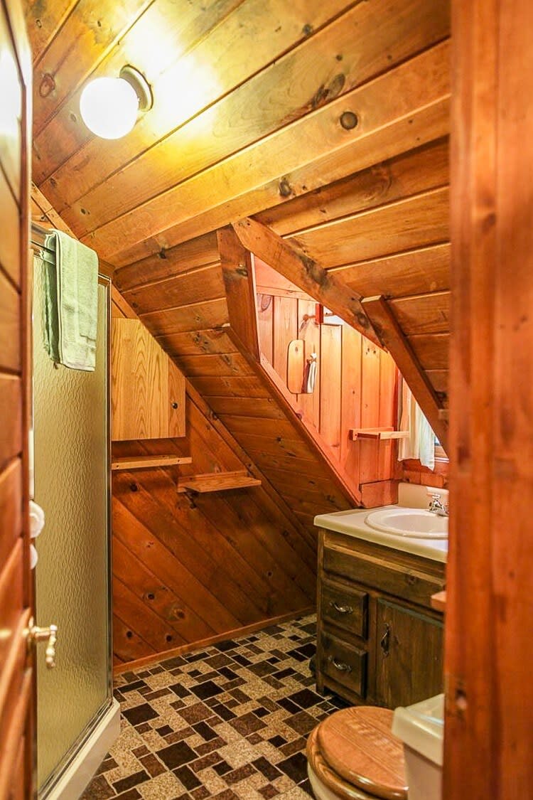 A rustic bathroom with sloped wooden plank walls and ceiling, featuring a shower stall, a vanity with a white sink, and a toilet with a wooden seat. The floor has brown and beige patterned tiles.