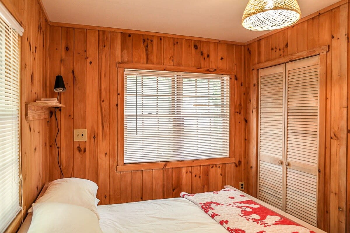 A corner of a rustic bedroom with knotty pine walls, showing a bed with white linens and a red quilt, two windows with blinds, and a louvered bi-fold closet door.