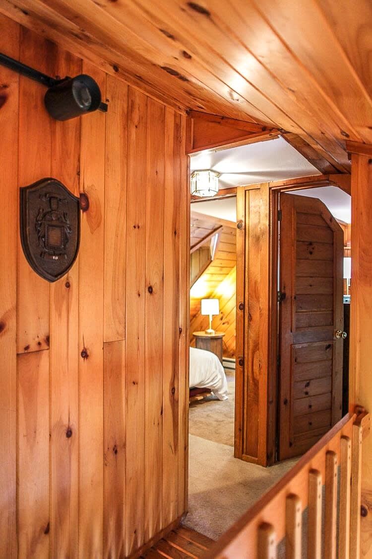 An upstairs hallway with knotty pine walls and ceiling, featuring a decorative wall plaque and leading to a bedroom with an open wooden door, revealing a glimpse of a bed and lamp.