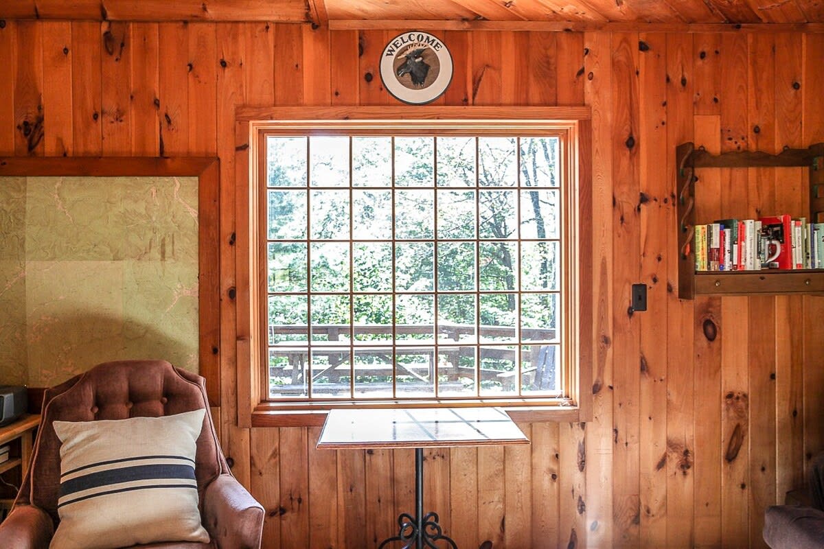 A bright living space with knotty pine walls, featuring a large grid-paned window offering a lush view of trees outside, a comfortable armchair with a striped pillow, and a small round table.
