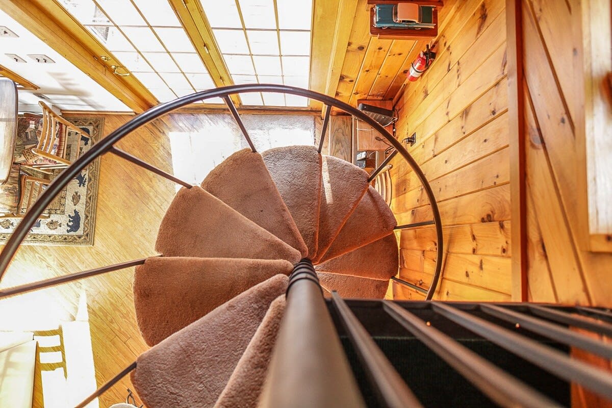 A bird's-eye view looking down the black metal spiral staircase with brown carpeted treads, showcasing the circular design and the warm knotty pine walls of the cabin's interior.