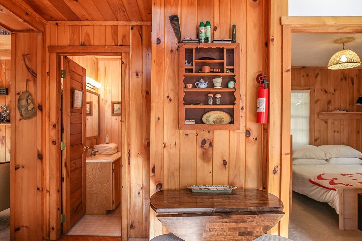 A rustic interior with knotty pine paneling throughout, featuring a small drop-leaf table, a wall-mounted shelf with decorative items, and a fire extinguisher, with glimpses into a bathroom and a bedroom.