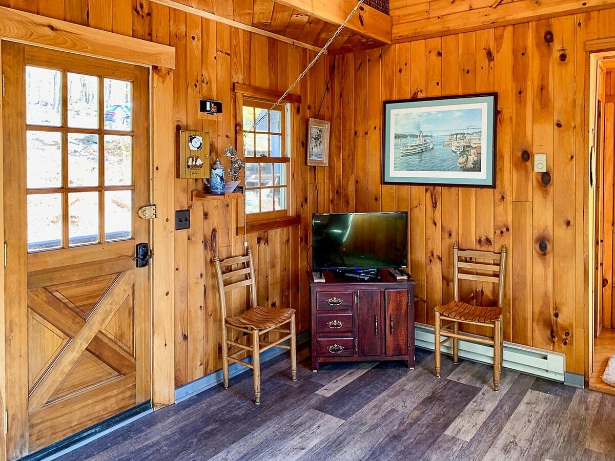 A section of the wood-paneled living room features a dark wooden TV stand with a television, flanked by two rustic wooden chairs, and a wooden door with glass panes leading outside.
