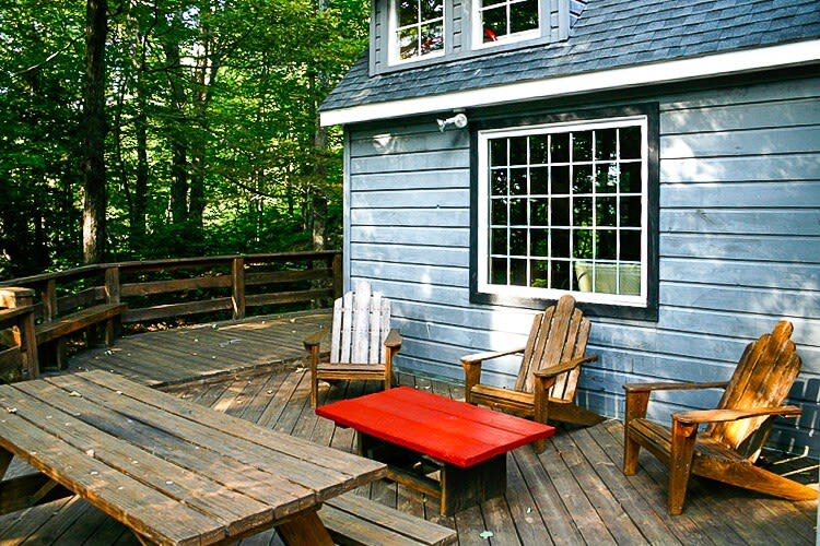 A spacious wooden deck with a picnic table, a red coffee table, and several Adirondack chairs, set against the backdrop of a gray-sided cabin and lush green trees. The deck features a wooden railing.