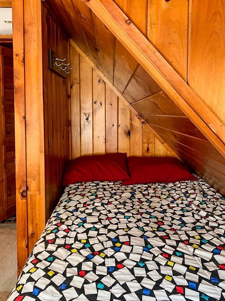 A cozy bedroom nestled under a sloped wooden ceiling, featuring a bed with a colorful geometric patterned comforter and two red pillows. A small coat rack is mounted on the wooden plank wall.