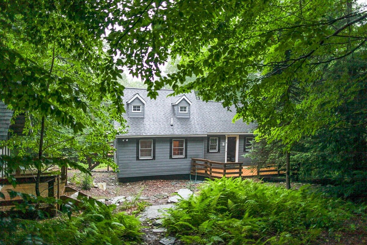 The back of the gray-sided cabin is visible through dense green foliage, featuring a small wooden deck and stairs, with dormer windows on the roof, blending into the natural landscape.