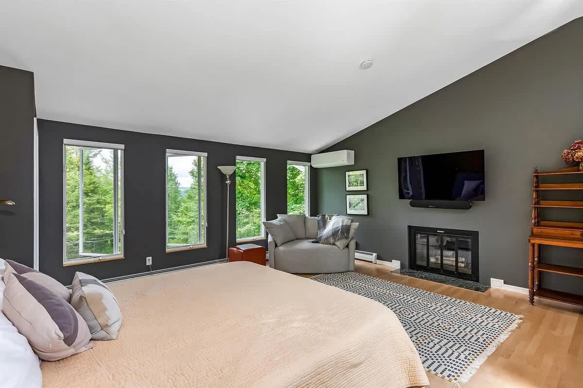 Another view of the spacious bedroom, highlighting the dark gray accent wall, a cozy armchair, and a fireplace with a TV mounted above. The room is filled with natural light from a series of tall windows.