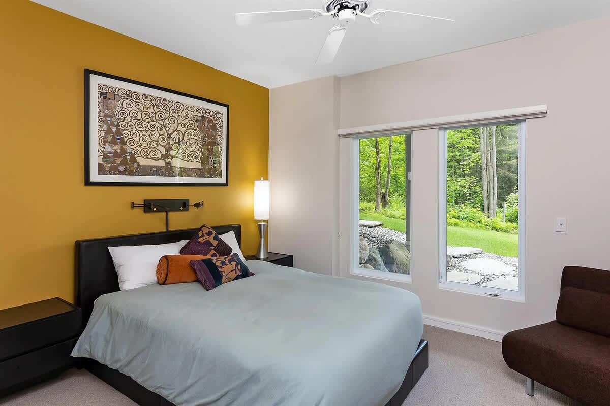 A modern bedroom featuring a queen-sized bed with a black headboard, a vibrant mustard yellow accent wall with a large framed artwork, and large windows offering a view of lush greenery.