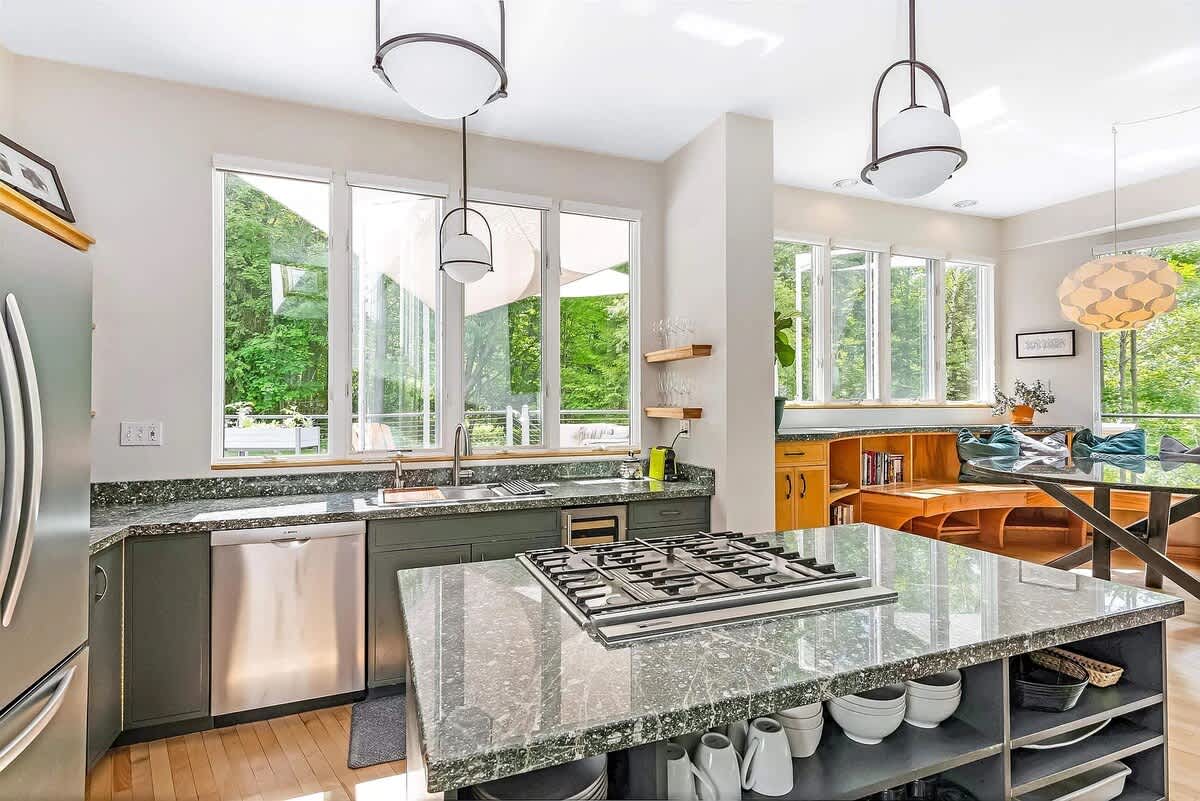 Contemporary kitchen with dark green cabinetry, speckled granite countertops, and a large island featuring a gas range. Large windows overlook the green outdoors, and two modern pendant lights illuminate the space.