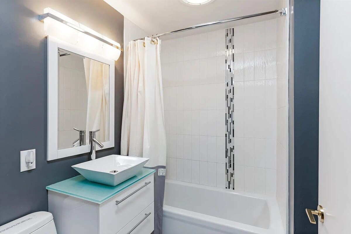 A modern bathroom with a white bathtub and shower combo, featuring white subway tiles with a decorative vertical mosaic stripe, a vessel sink on a white vanity, and a large mirror.