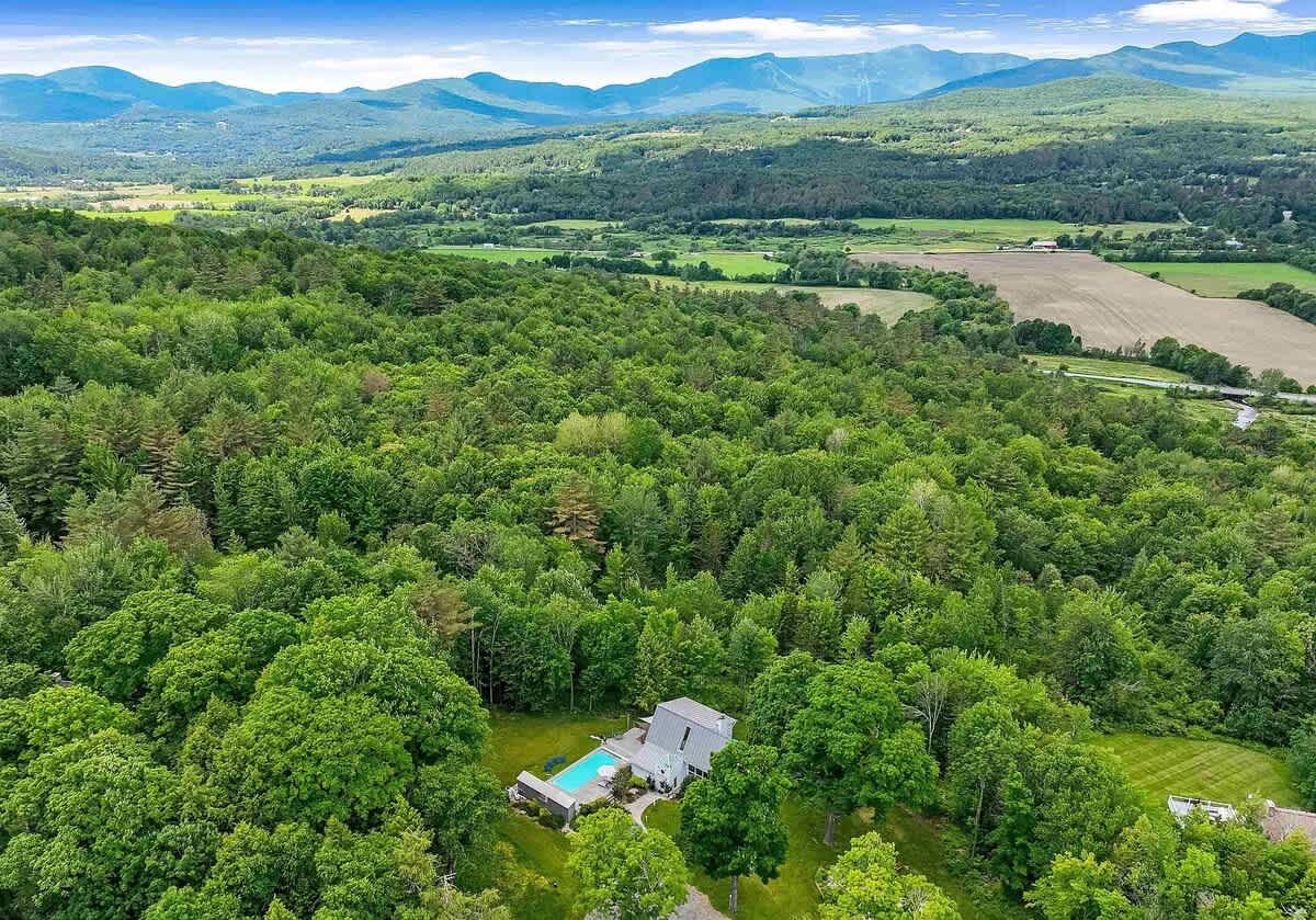 Aerial view of a modern house with a swimming pool, nestled within a dense green forest, with rolling hills and distant mountains under a clear blue sky.