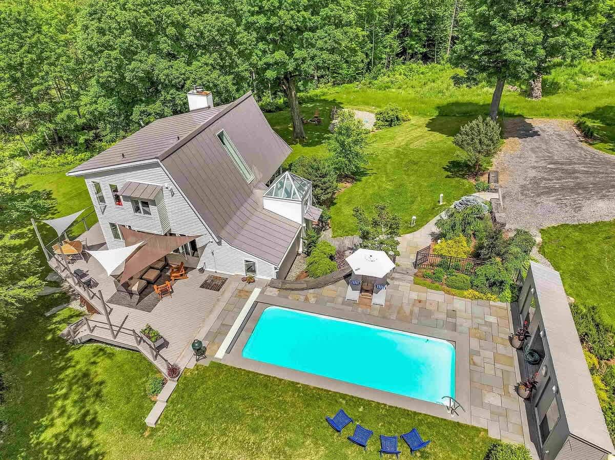 Aerial view of a modern house with a gray roof, surrounded by lush green trees and lawn. The property features a rectangular swimming pool with a stone patio, multiple outdoor seating areas, and a long driveway.