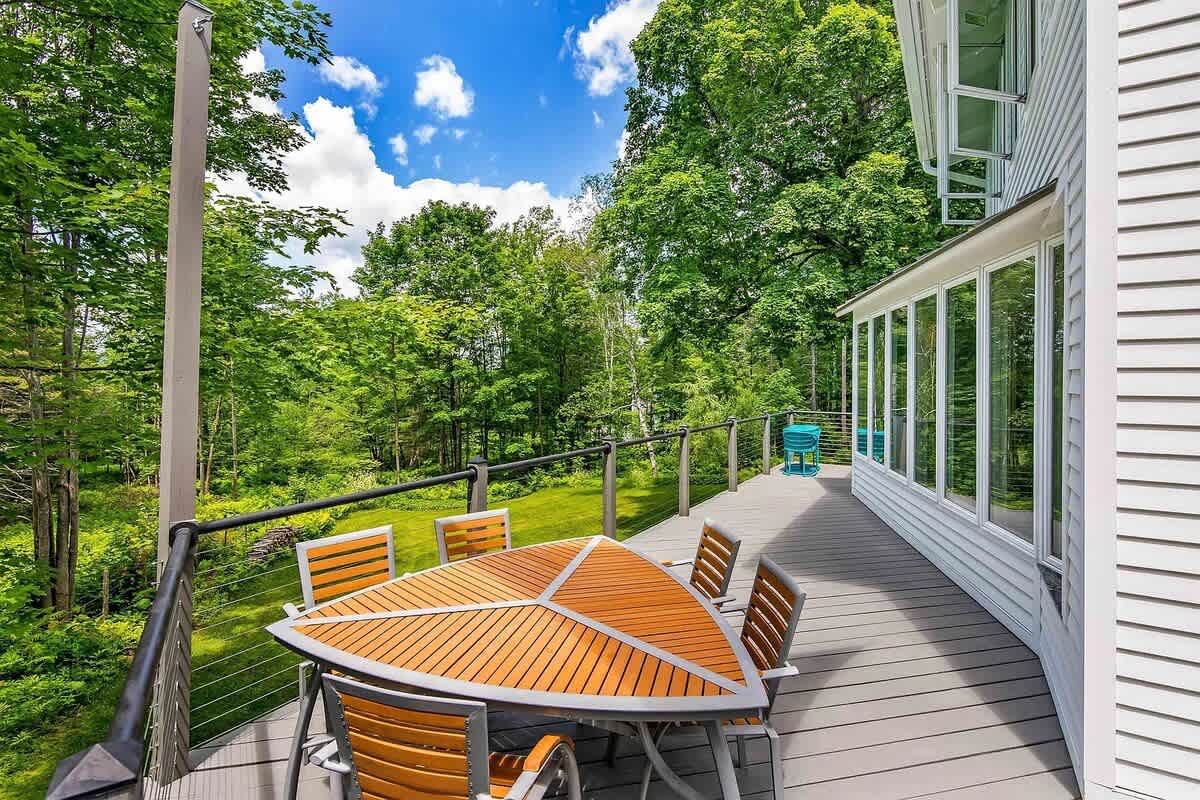 A triangular outdoor dining table with matching chairs on a spacious deck, offering panoramic views of the surrounding vibrant green forest under a bright blue sky.