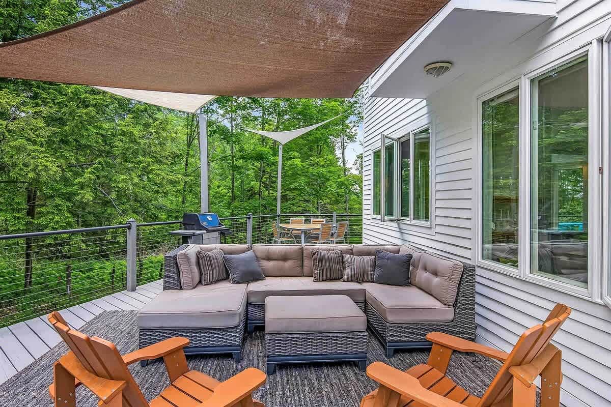 A spacious outdoor deck featuring a comfortable sectional sofa with an ottoman under a large shade sail, and two wooden Adirondack chairs, all overlooking a lush wooded area.