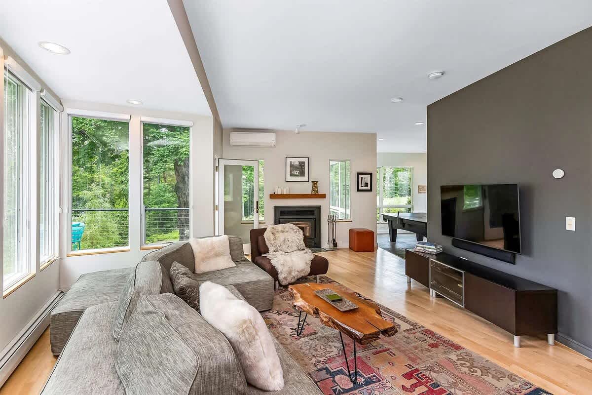 Spacious living room with a grey sectional sofa, a unique wood coffee table, and a wall-mounted TV, connected to a game room with a ping pong table, all featuring large windows and hardwood floors.
