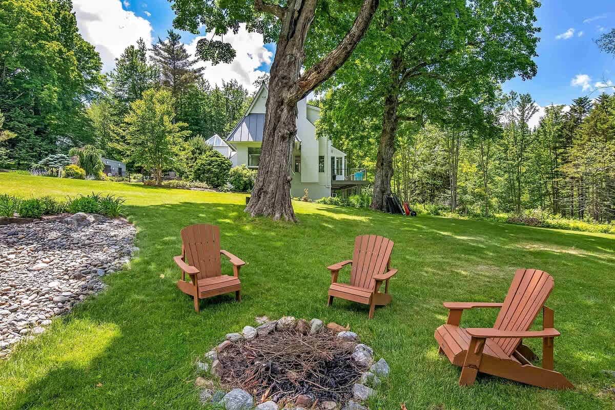 A cozy outdoor fire pit area with three wooden Adirondack chairs arranged around a stone-lined fire pit, set on a manicured lawn with large mature trees and the house in the background.