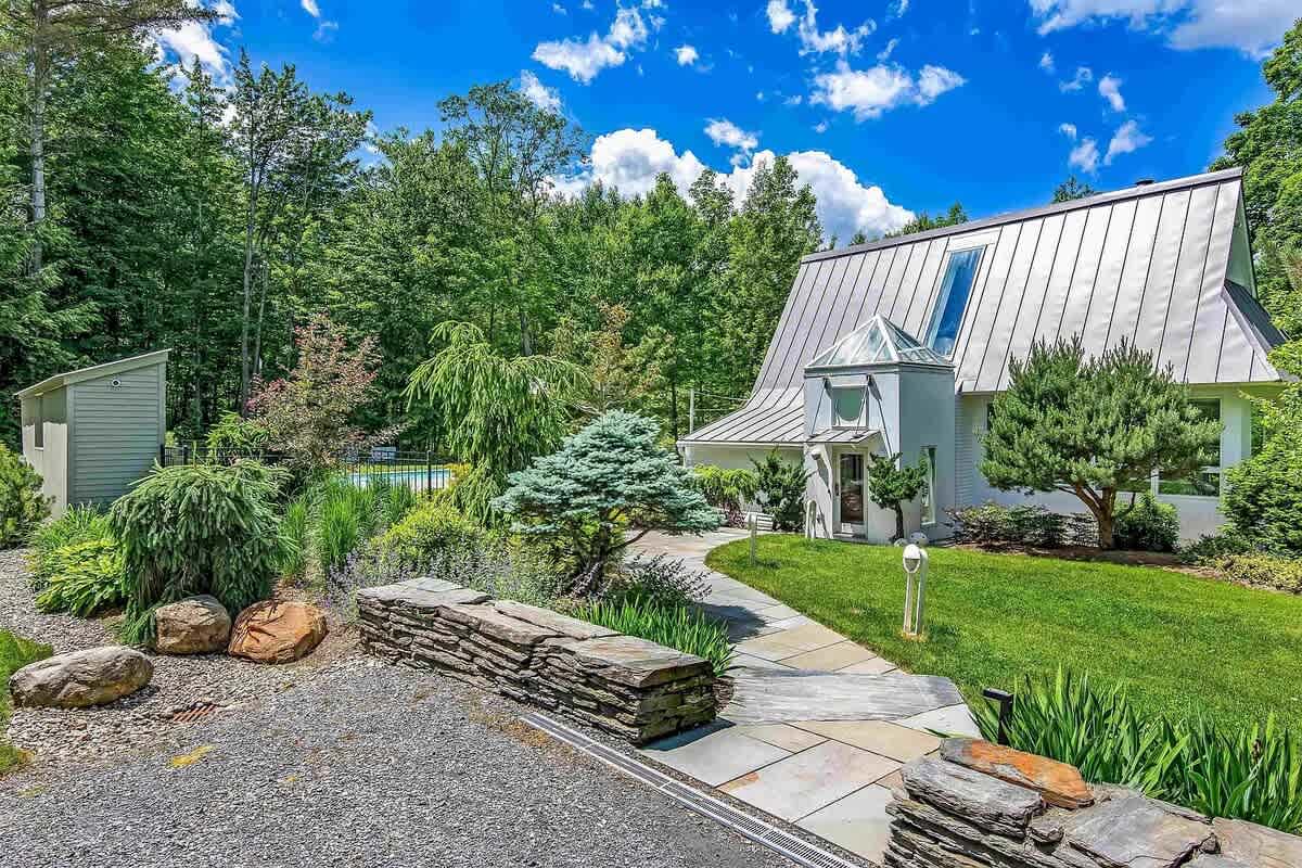 Modern white house with a striking metal roof and a unique entrance, surrounded by lush green landscaping, a stone pathway, and a small shed, under a bright summer sky.