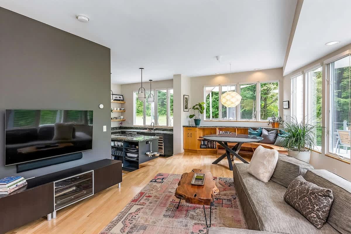 Open-concept living room with light hardwood floors, a large grey sectional sofa, a unique live-edge coffee table, and a wall-mounted TV, flowing into a kitchen area with large windows.