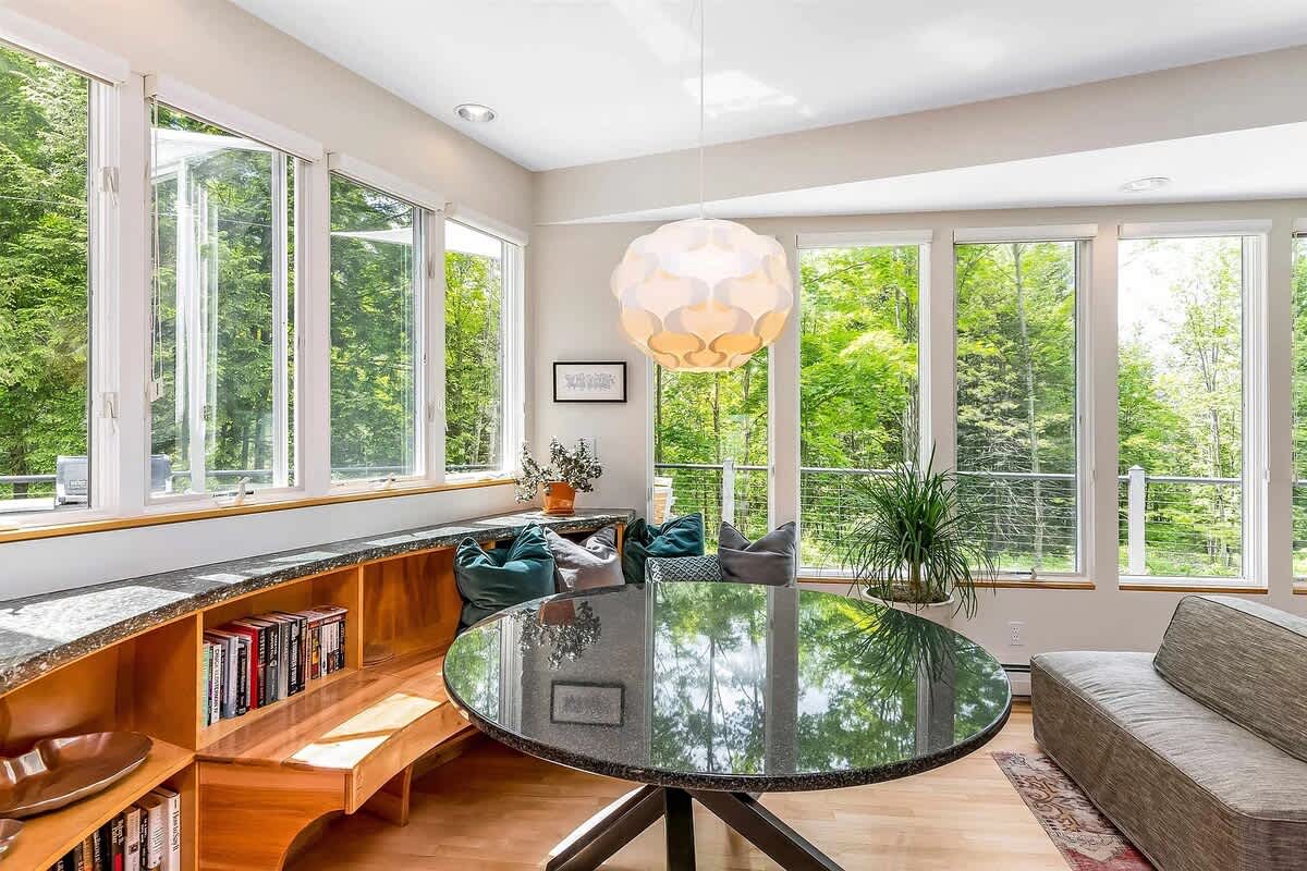 Modern dining area featuring a round black granite table, built-in wooden bench seating with bookshelves, and large windows offering lush green outdoor views. A unique spherical pendant light hangs above the table.